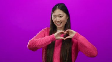 Pembe kazak giymiş, elleri ile kalp şeklinde gülümseyen genç, Asyalı, dışlanmış, dışa dönük, yetişkin, oyuncu bir kadının yer aldığı canlı pembe bir duvara karşı sevgi ve pozitiflik taşıyan bir kadın..