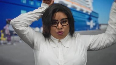 Genç İspanyol kadın deniz kıyısındaki bir limanda, büyük bir teknenin önünde elleriyle kulaklarını taklit ederek şakacı hareketler yapıyor..