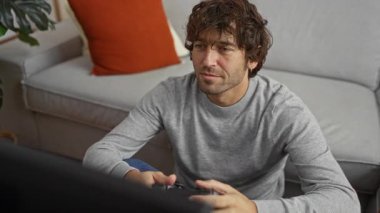 Genç adam rahat bir oturma odasında kanepede oturuyor, bir kontrolörle video oyunları oynamaya odaklanmış, rahat bir ev ortamında keyif ve nişan sergiliyor..