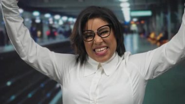 Tren istasyonunda kendinden emin bir şekilde gülümseyen, kendini gösteren başparmaklarıyla işaret eden, gözlük ve beyaz gömlek takan, kapalı alanda kendini ifade etme gücünü yakalayan bir kadın..