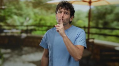 İspanyol adam, güneşli bir restoranın terasında sessiz kalmak için jest yapıyor yemyeşil bir ortam yaratıyor..