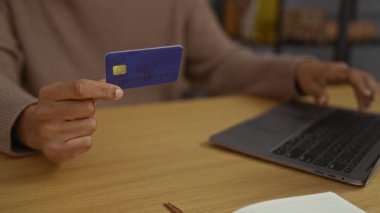 Bilgisayarda çalışan genç adam, modern rahat çalışma alanında kredi kartı tutuyor ve online alışveriş veya dijital ödeme yapıyor.