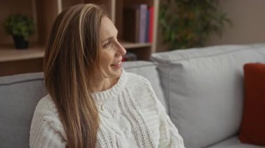 Oturma odasındaki rahat koltukta beyaz kazak giyen, sarı saçlı, kapalı koltukta gülümseyen kadın mutluluk ve rahatlama gösteriyor..