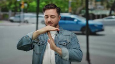 Sakallı genç adam şehir sokaklarında el hareketi yapıyor ve şehir ortamında gündüz vakti arka planda arabalar bulanıklaşıyor.