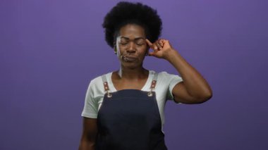 Önlük takan genç bir kadın, parlak mor bir arkaplan karşısında başını kapalı tutarak kafasını işaret ediyor, kafa karışıklığı veya düşünceli bir ifadeyle münferit bir ifade sergiliyor..