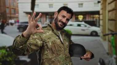 İspanyol üniformalı bir adam kalabalık bir sokakta elinde miğfer tutarak gülümsüyor. Bu da kendine güveni ve açık havada pozitif tutumunu gösteriyor..