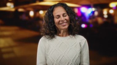 Açık havada gülümseyen beyaz kazak giyen, kıvırcık saçlı bir kadın. Şehir atmosferini aydınlatıyor..