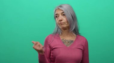 Gri saçlı yaşlı kadın, yeşil arka plan duvarına karşı, göğsünde dövmesi olan pembe gömlekli, kendine güvenini ve tarzını gösteriyor..