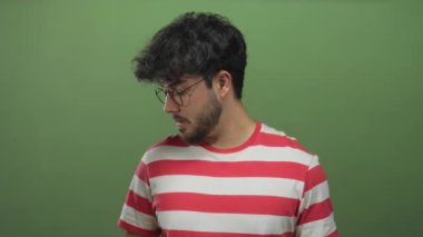 Çizgili tişörtlü, sakallı ve gözlüklü genç bir İspanyol stüdyo ortamında yeşil arka plana karşı şaşırmış görünüyor..