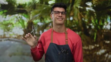 Parkta gözlüklü ve önlüklü genç İspanyol bir adam, kendinden emin bir şekilde bir kuaförün karizmasını betimliyor açık hava yeşilliğinin ortasında, modern stil ve beceri yayıyor..