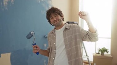 Genç adam duvarları parlak, yeni oturma odasında boyuyor, gülümsüyor ve gevşek bir jestle boya yuvarlıyor, ev dekorasyonunda bir neşe ve başarı duygusunu somutlaştırıyor..