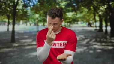 Kırmızı gömlekli genç adam dışarıda parkta jest yapıyor. Arka planda ağaçlar var. Davetkar bir jest yaparken neşeli bir ifade sergiliyor..