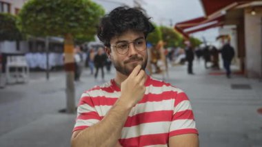 Çizgili gömlekli, sakallı ve gözlüklü genç adam bulanık arka planı ve ağaçları olan yoğun bir sokakta dalgın dalgın duruyor..