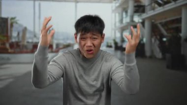 Şehir sokaklarında duygusal olarak el kol hareketi yapan genç Çinli bir adam, gri bir kazak giyiyor, arka planda modern şehir mimarisi var..