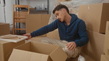 Yeni dairede telefonla konuşurken kutuları açan genç adam, kartonla çevrili, günlük kıyafetler giyiyor, heyecan gösteriyor, şehirdeki modern yaşam tarzını yansıtıyor..