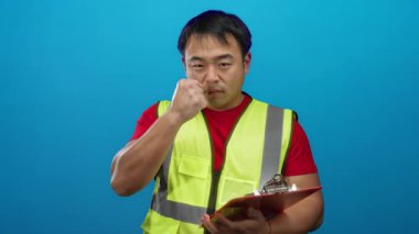 Emniyet yeleği giyen bir adam mavi arkaplan üzerinde pano tutarken tanımlı bir ifade gösteriyor.