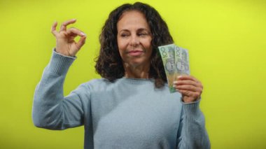 Avustralya doları tutan orta yaşlı bir kadın canlı sarı bir duvara karşı güvenli bir şekilde meditasyon yapıyor, sakin finansal başarı ve farkındalığı somutlaştırıyor..