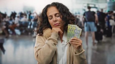 Romanyalı kadın, yoğun bir havaalanı terminalinde kapalı bir odada derin düşüncelere dalmış ve hareketli bir seyahat düzeninde düşünceli görünüyor..