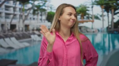Açık havuz kenarında hiç havuzu olmayan, pembe tişörtlü, el işaretli reddeden, güneşli atmosferli, çevresi otel ve palmiye ağaçlarıyla çevrili bir kadın..
