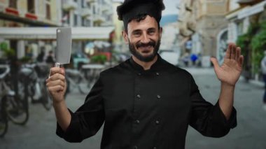 Sakallı, siyah üniformalı İspanyol aşçı balta tutuyor açık havada, hareketli bir sokakta gülümsüyor..