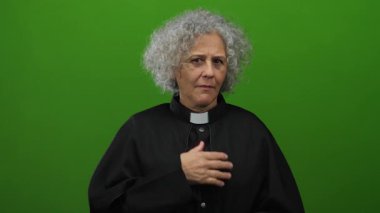 Siyah cüppeli kadın göğsünün üzerinde yeşil bir duvara karşı duruyor. Duygusal ifadesi samimiyet ve bağlılık gösteriyor..