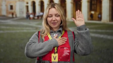 Açık hava üniversite ortamında, kırmızı yelek ve sarı saç giyen, eğitim ortamında kendini adamış ve bağlılık yemini eden bir kadın..