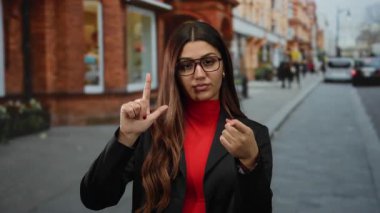 Gözlük takan kadın, şehir sokağında arka planda kırmızı tuğlalı binalarla ezik jestler yapıyor. Dışarıdaki hareketli ortamda kendine güvenen tavırlar sergiliyor..