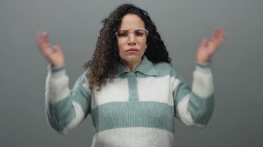 Kıvırcık saçlı ve gözlüklü bir kadın elleriyle kulaklarını kapatıyor, gri bir arka plana karşı stres ifade ediyor, çizgili bir kazak giyiyor endişeli bir ifadeyle, kapalı alanda izole edilmiş..