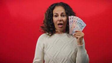 Orta yaşlı bir kadın arkasında kırmızı bir geçmişle Thai banyosu parası tutarken şaşırdı. Finansal başarı ya da talih kuşu gibi canlı bir ifade ve renkli notlar sergiliyordu..