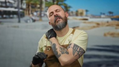 Kel, sakallı ve dövmeli bir adam deniz kenarındaki güneşli bir güvertede boynuna masaj yapıyor ve deniz kenarında rahatsız olduğunu söylüyor..