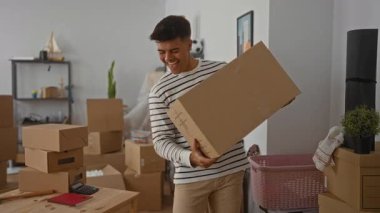 Genç adam yeni bir apartman dairesinde kutuları açarken gülümsüyor modern oturma odasını gösteriyor ve dağınık karton kutuların arasında heyecan yaratıyor..
