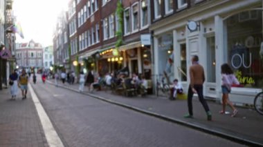 Amsterdam 'daki bulanık şehir caddesinde yürüyen insanlar, her iki taraftaki binalar ve sıcak akşam ışığı, canlı ama huzurlu şehir atmosferini yansıtıyor..