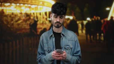 Gece fuarında akıllı telefon kullanan genç adam parlak ışıklar ve arka planda sıradan kot ceket giyen insanlar ile düşünceli görünüyor..