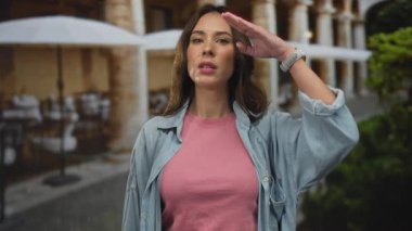 Sıradan bir kot ceketle asker selamı veren genç bir kadın hayat dolu bir şehirde sokak manzaralı ve açık hava manzaralı bir restoran terasında duruyor..