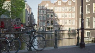 Bisiklet tırabzana yaslanmış, arka planda amfi kanalları ve geleneksel Hollanda binaları, yeşillik ve güneş ışığıyla çevrili, sakin bir sahne yaratıyor..