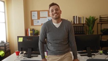 Bilgisayarlar ve kitaplarla dolu modern ofis ortamında güvenle gülümseyen genç bir adam profesyonel ve ulaşılabilir bir atmosfer yaratıyor..