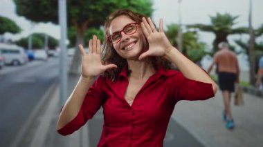 Kırmızı tişörtlü, gözlüklü bir kadın şehir sokağında duruyor. Arka planda bulanık insanlar ve yeşillik var. Mutluluk saçıyor ve kaygısız bir hava var..