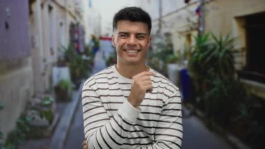 Neşeli bir şehir sokağında, çizgili bir tişört giymiş, güneşli bir günde, yemyeşil bir sokaktaki çiçeklerle çevrili, kendine güvenen genç bir adam..