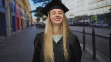 Şehir sokaklarında mezuniyeti kutlayan kadın, kep ve cübbe giyiyor, heyecanla ve gururla gülümsüyor..