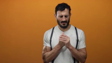 Sakallı İspanyol adam canlı turuncu arka planda duruyor, beyaz gömlek ve pantolon askısı giyiyor, çeşitli düşünceli ifadeler sergiliyor..