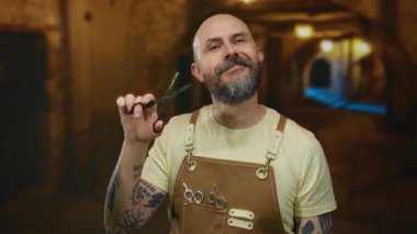 Sakallı, dövmeli, sarı tişörtlü, kahverengi önlüklü, elinde makas tutan, arka planda bulanık ışıklar olan kel bir adam..