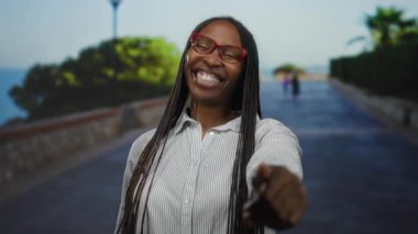 Deniz kenarında bir gezinti alanında gözlüklü bir kadın, güneşli bir plaj gününde neşeli bir ifade sergiliyor..