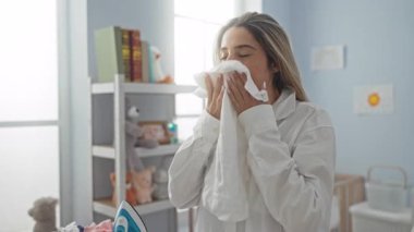 Parlak bir çamaşırhanede yeni ütülenmiş kıyafetlerden hoşlanan bir kadın, rahat bir gülümsemeyle etrafı ev eşyaları ve gün ışığıyla çevrili beyaz bir gömlek giyerken..