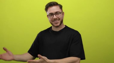 Gözlüklü genç adam parlak sarı bir arka plana karşı ellerini açık sunuyor neşeli ve çekici görünüyor, iş ve yaratıcı sunumlar için ideal..