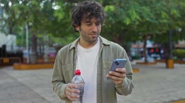 Parkta elinde telefon ve su şişesi olan bir adam perdeye bakıyor, günlük kıyafetler, şehir manzarası, güneşli bir gün, gevşemiş bir ruh hali..