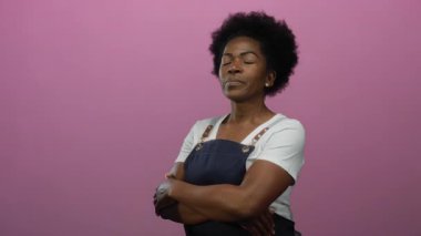 Önlük takan, parlak pembe arka planda gülümseyen, çapraz kollu, kendine güvenen, ince, izole, duygusal, canlı modern bir kadın.