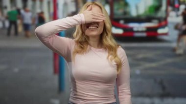 Kalabalık bir şehir caddesinde arka planda kırmızı bir otobüsle gülümseyen bir kadın, neşeli bir şekilde gözlerini kapatıyor, şehir hayatı ve hareketli bir trafik ile çevrili..