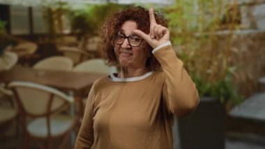 Kadın esmer orta yaşlı jestler açık hava restoranının terasında kaybedenler kahve ve sandalyelerle arka planda komik ve oyuncu ifadeleri resmediyor..