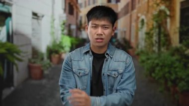 Genç Çinli bir adam canlı bir şehir caddesinde kot ceket giyerek jest yapıyor, açık hava ortamında sıradan bir şehir yaşam tarzını çağrıştırıyor..