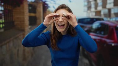 Mavi kazaklı genç bir kadın işlek bir şehir caddesinde dürbün taklidi yapıyor. Arka planda şehir binaları ve park halindeki arabalarla çevrili neşeli bir ifade var..